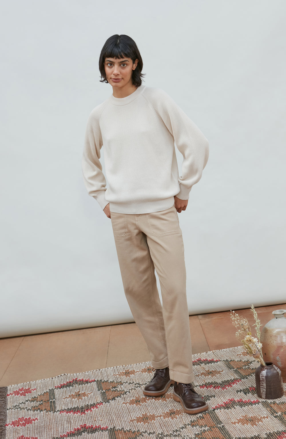 A person stands indoors wearing a cream crewneck cotton knitted rib sweatshirt, beige pants, and dark brown shoes, posed on a patterned rug against a plain light wall. Made by Paul James Knitwear.