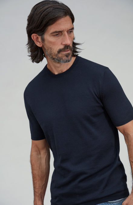 A man wearing a plain dark navy cotton short-sleeve crewneck t-shirt, photographed from the chest up against a light gray studio background. By Paul James Knitwear