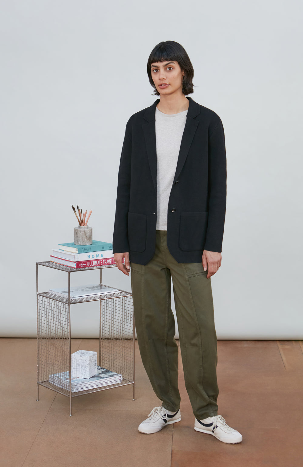 A model wearing a black knitted blazer by Paul James Knitwear over a light gray crewneck t-shirt and olive green pants, standing in a minimalist room with a white wall and a small wire shelf.