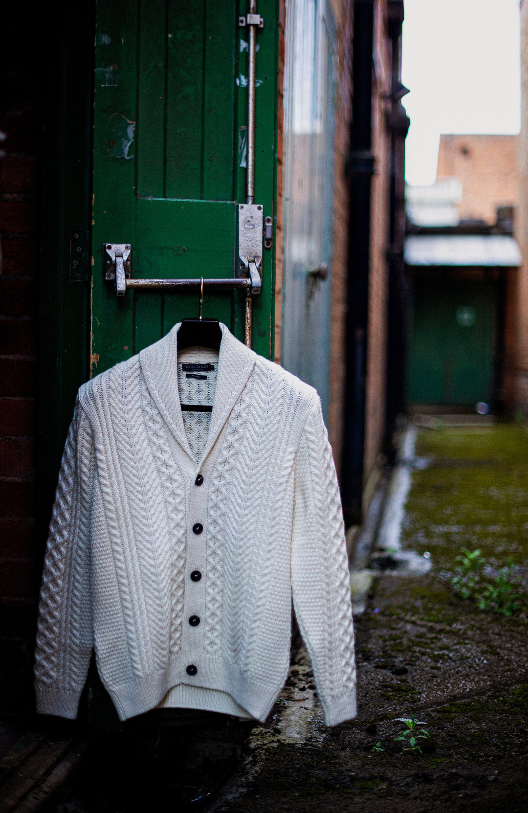 Men's made-in-England wool cardigan. A white cable-knit cardigan sweater hanging on a black hanger against a dark green wooden door in an outdoor alleyway.