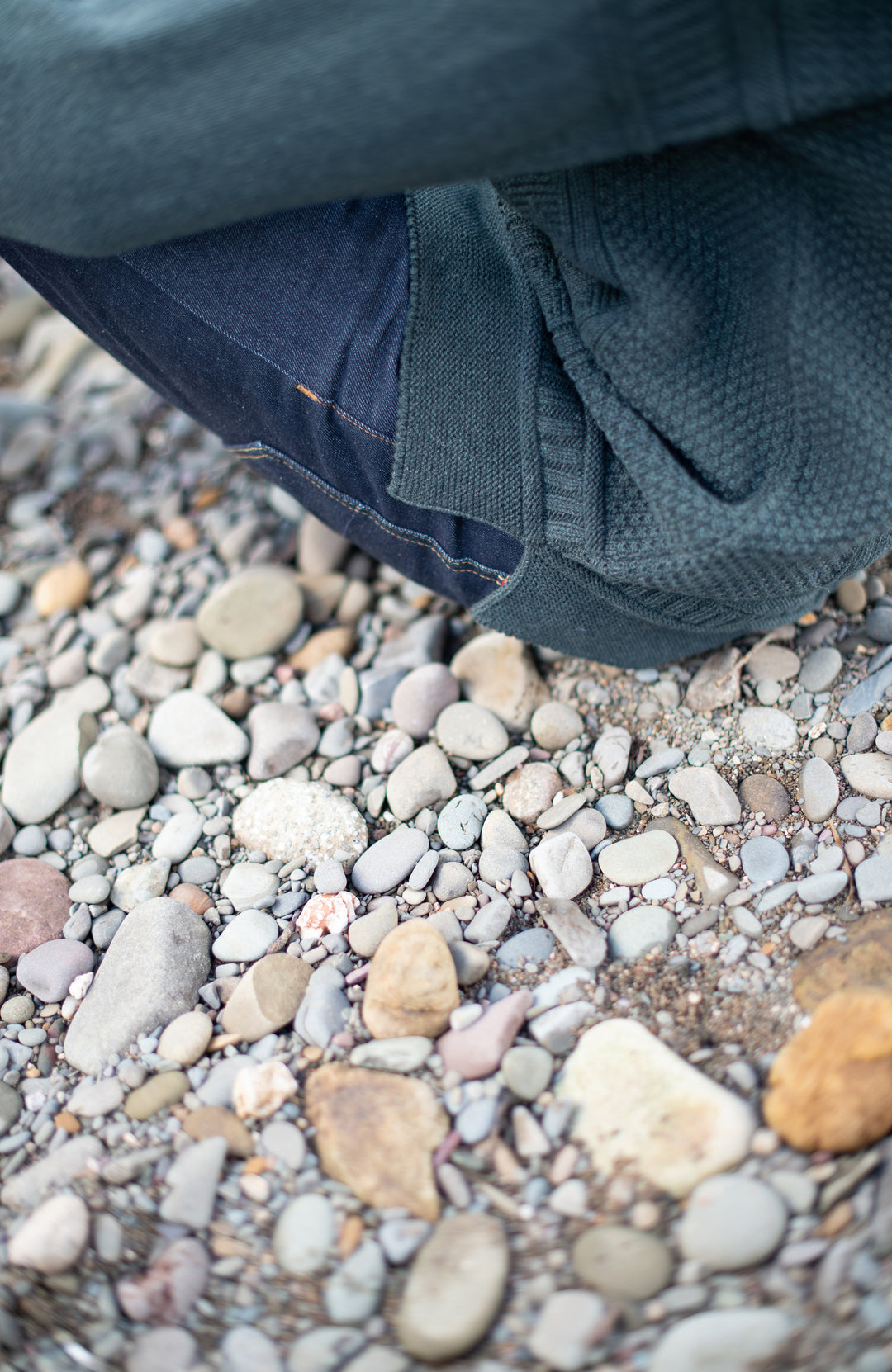 A close-up lifestyle shot of a person's legs and clothing resting on a bed of small, smooth river stones and gravel.