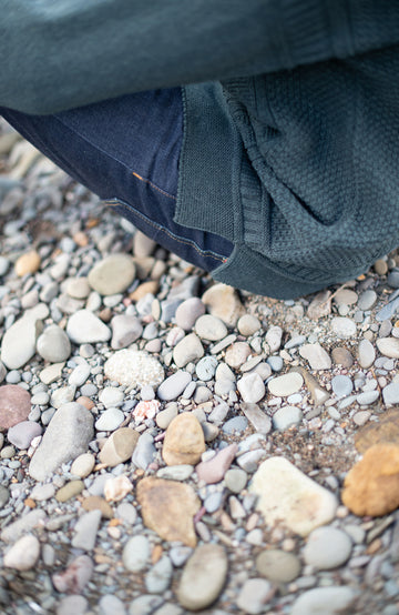 A close-up lifestyle shot of a person's legs and clothing resting on a bed of small, smooth river stones and gravel.