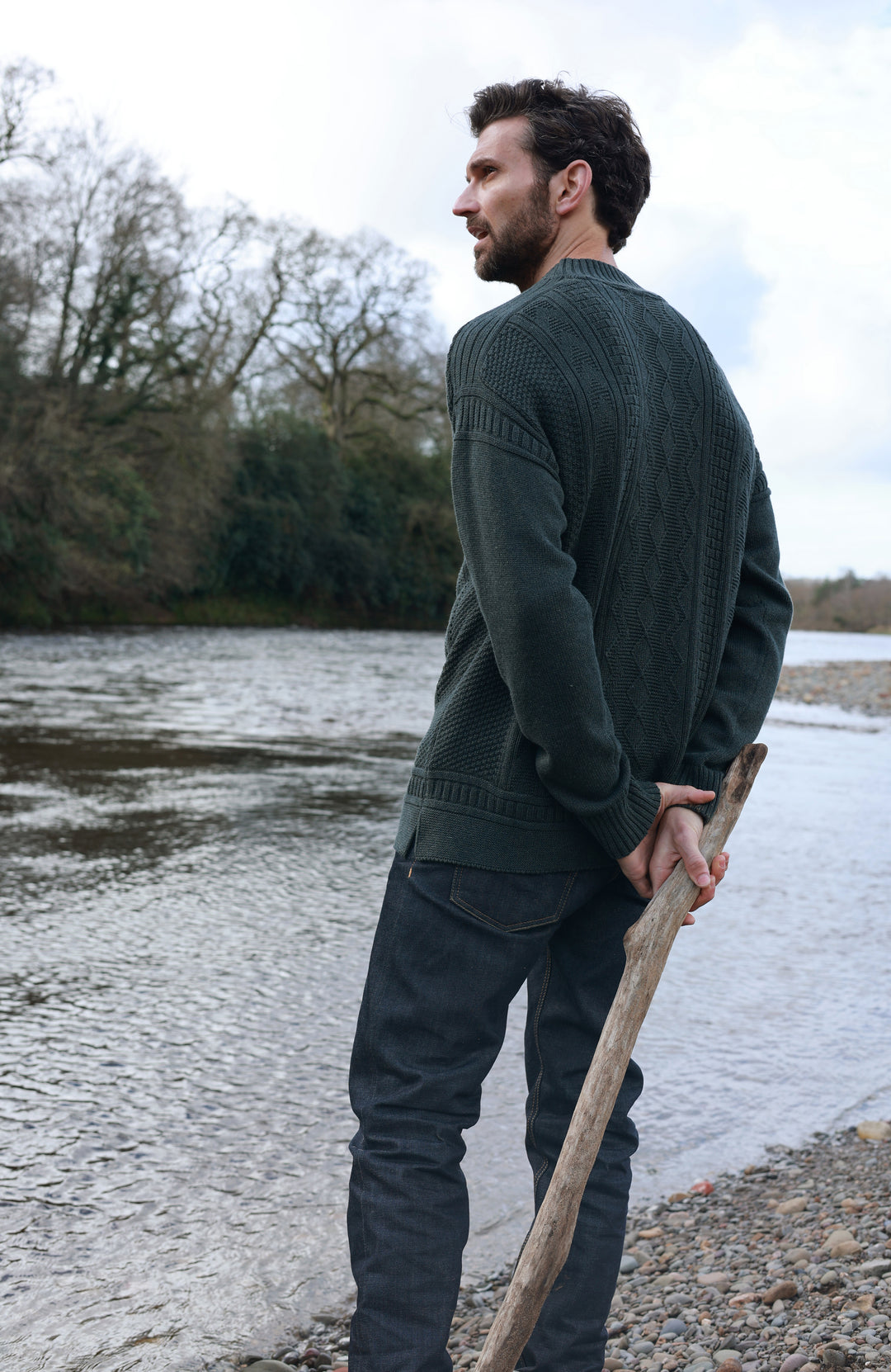 A man wearing a dark green ribbed knit mock neck guernsey sweater by Paul James Knitwea and dark jeans stands on a pebble shore next to a river, holding a wooden stick and looking off-camera.