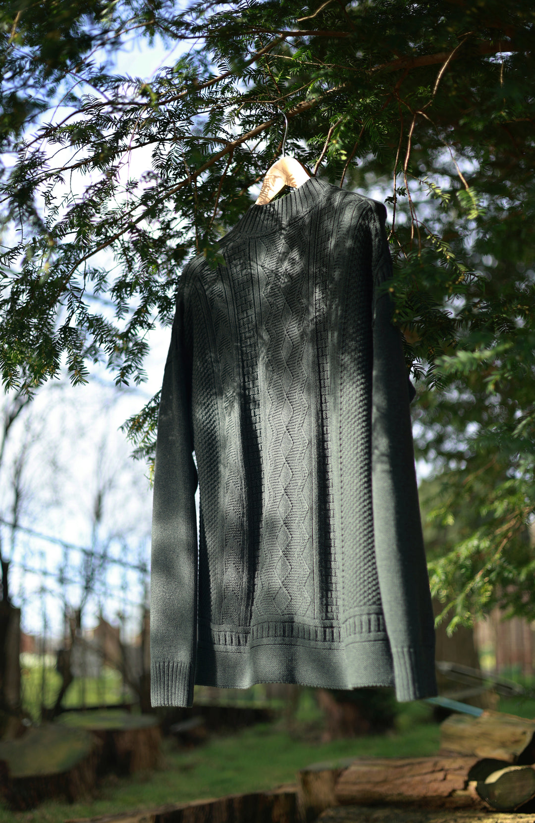 dark green ribbed knit mock neck guernsey sweater by Paul James Knitwea hanging from a wooden hanger on a tree branch outdoors, with green pine needles and a blurred natural background.