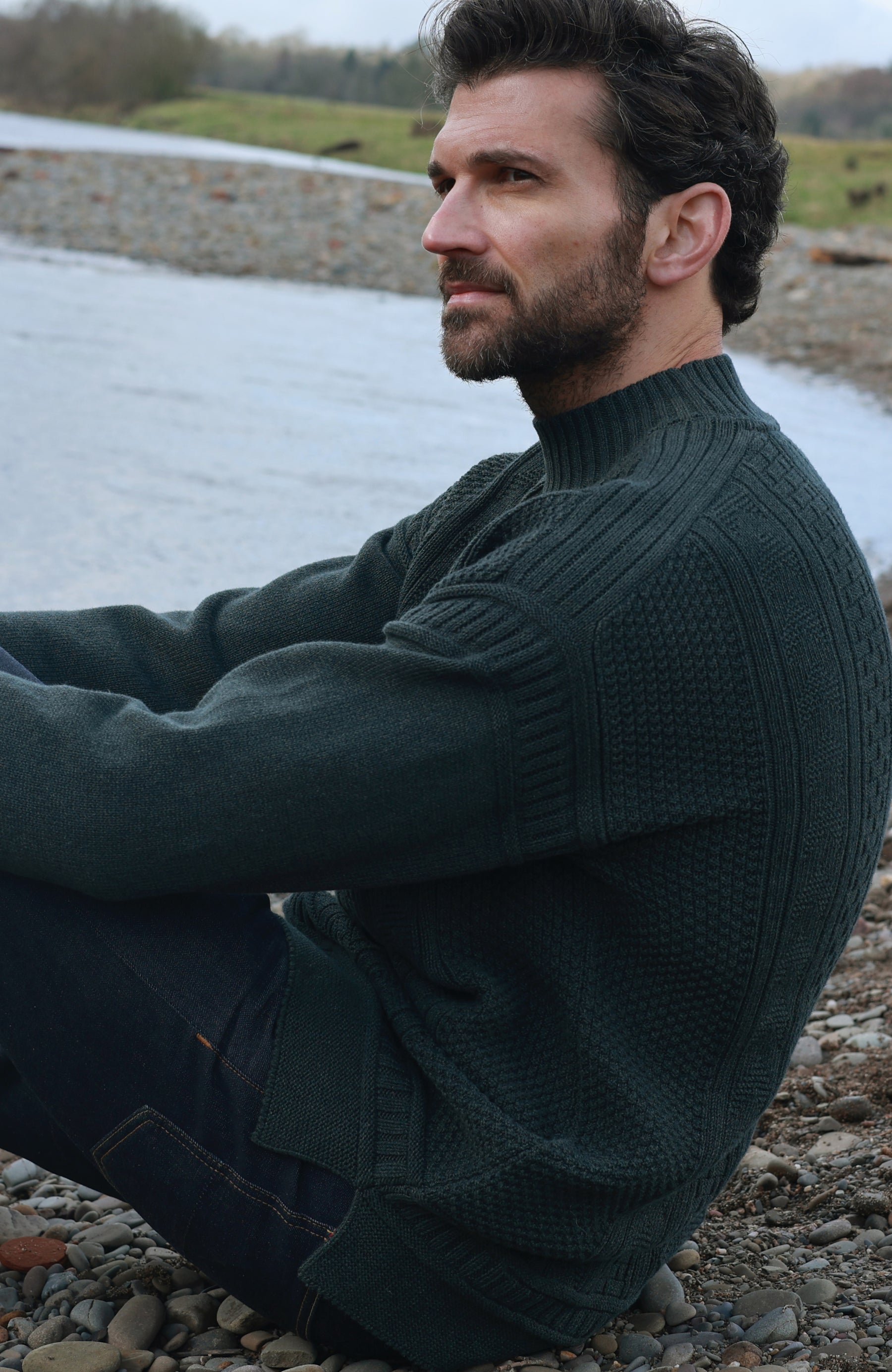A man wearing a dark green ribbed knit mock neck guernsey sweater by Paul James Knitwear sits on a pebble-strewn shore with water and grass in the background.