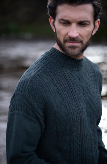 A man wearing a dark green ribbed knit mock neck guernsey sweater by Paul James Knitwea, photographed outdoors with a blurred natural background.