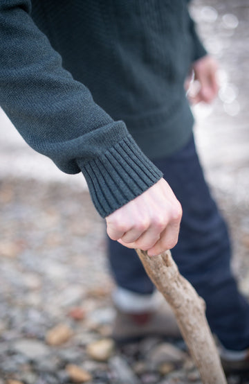 A close-up of a person outdoors holding a rough wooden stick or walking cane, wearing a dark ribbed knit sweater and dark pants on a pebble-covered ground.