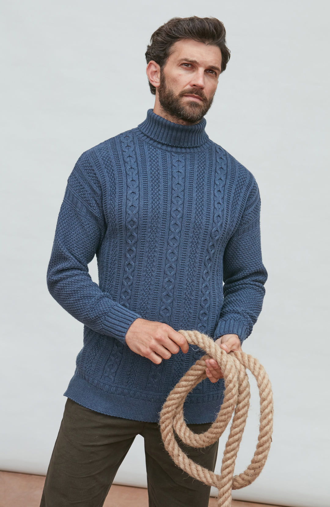 Man wearing a blue cable knit sweater holding a rope against a light background