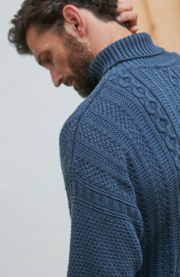 Man wearing a blue paul james turtle neck cable sweater against a light background