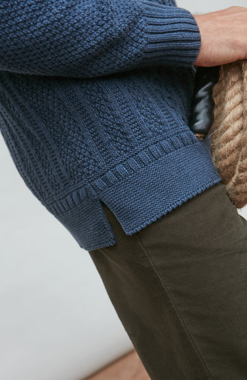 Person wearing a blue paul james turtle neck cable sweater and brown pants on a white background
