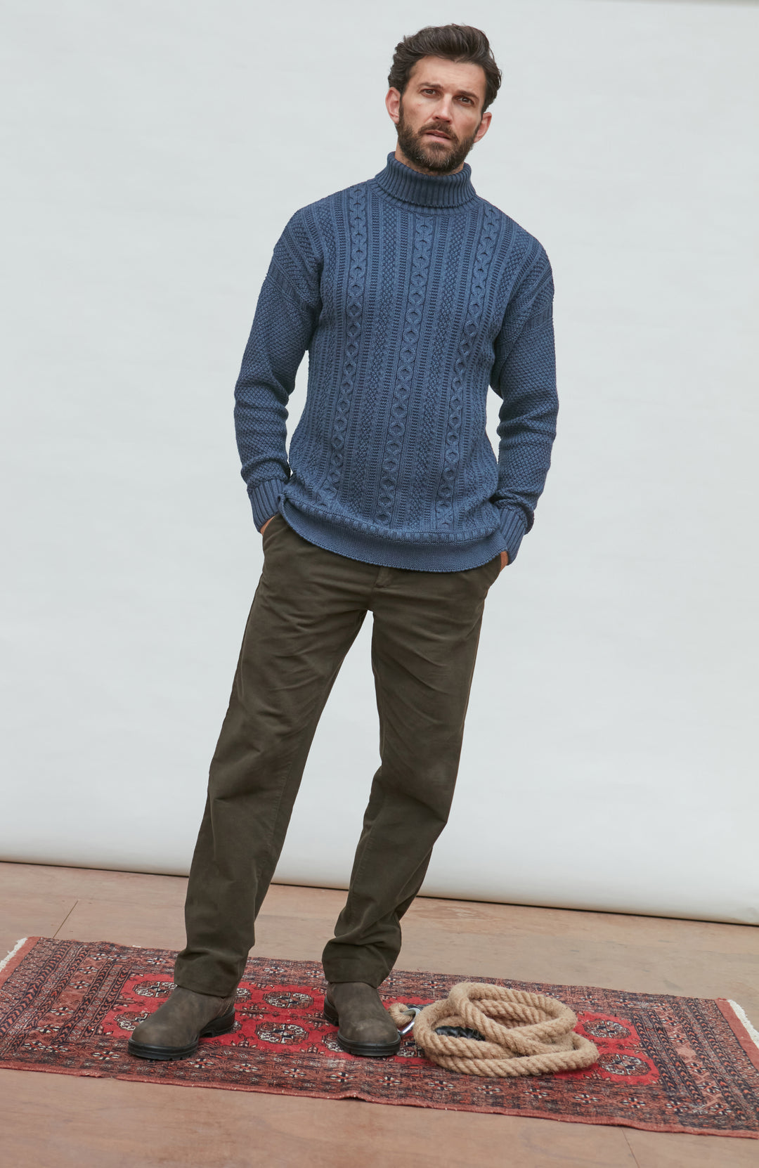 Man wearing a blue paul james turtle neck cable sweater and green pants standing on a wooden floor with a white wall background.