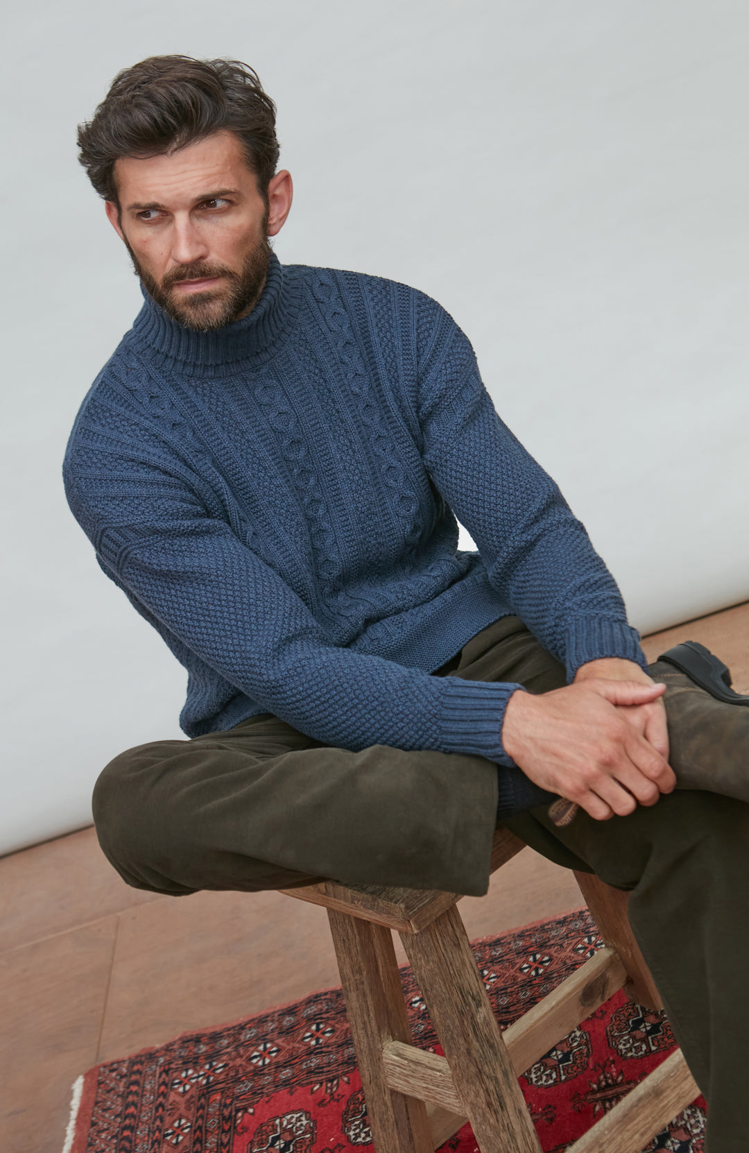 Man wearing a blue paul james turtle neck cable gansey sitting on a wooden stool with a white wall background