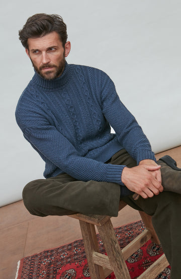 Man wearing a blue paul james turtle neck cable gansey sitting on a wooden stool with a white wall background