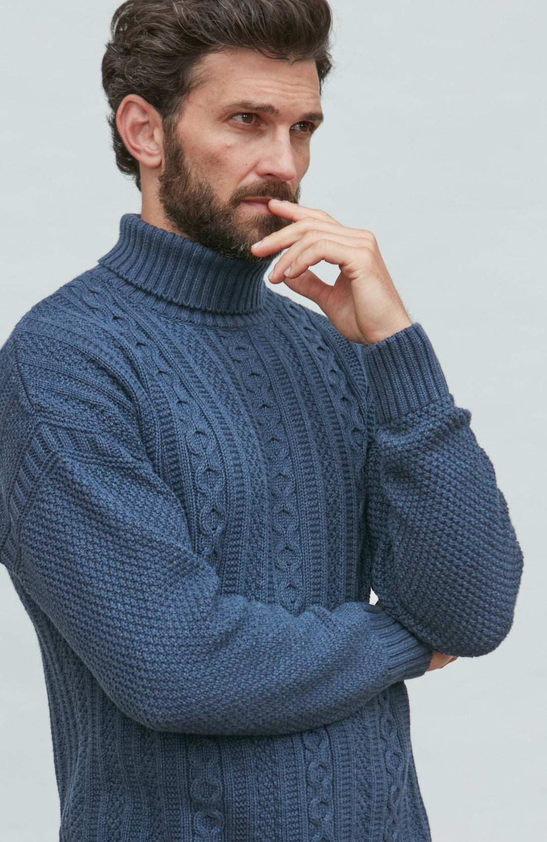 Man wearing a blue paul james turtle neck cable guernsey against a light gray background