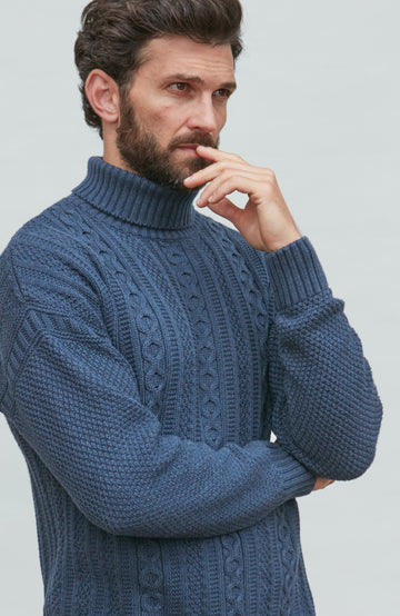 Man wearing a blue paul james turtle neck cable guernsey against a light gray background