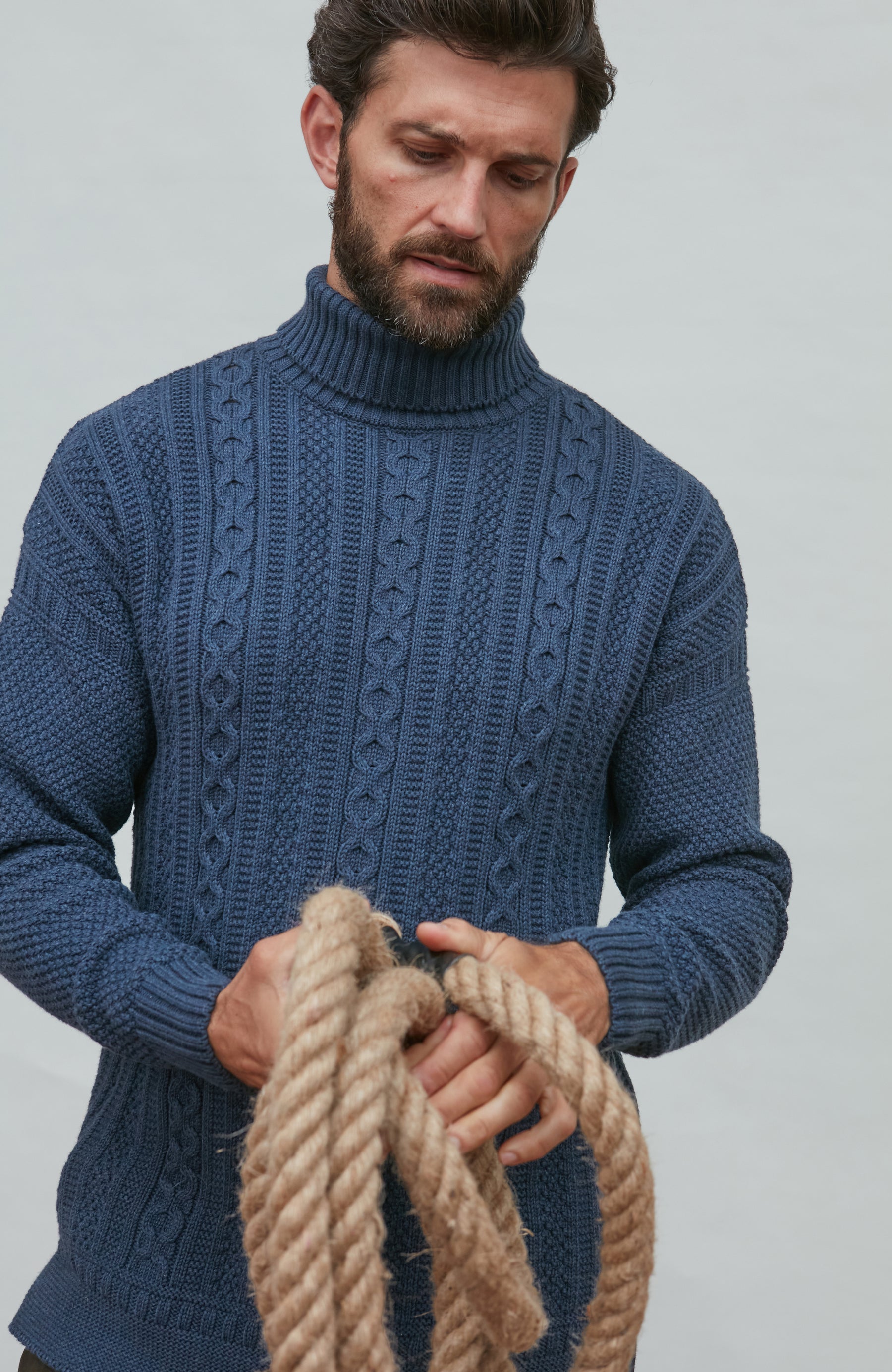 Man wearing a blue paul james turtle neck cable knitted sweater holding rope against a plain background
