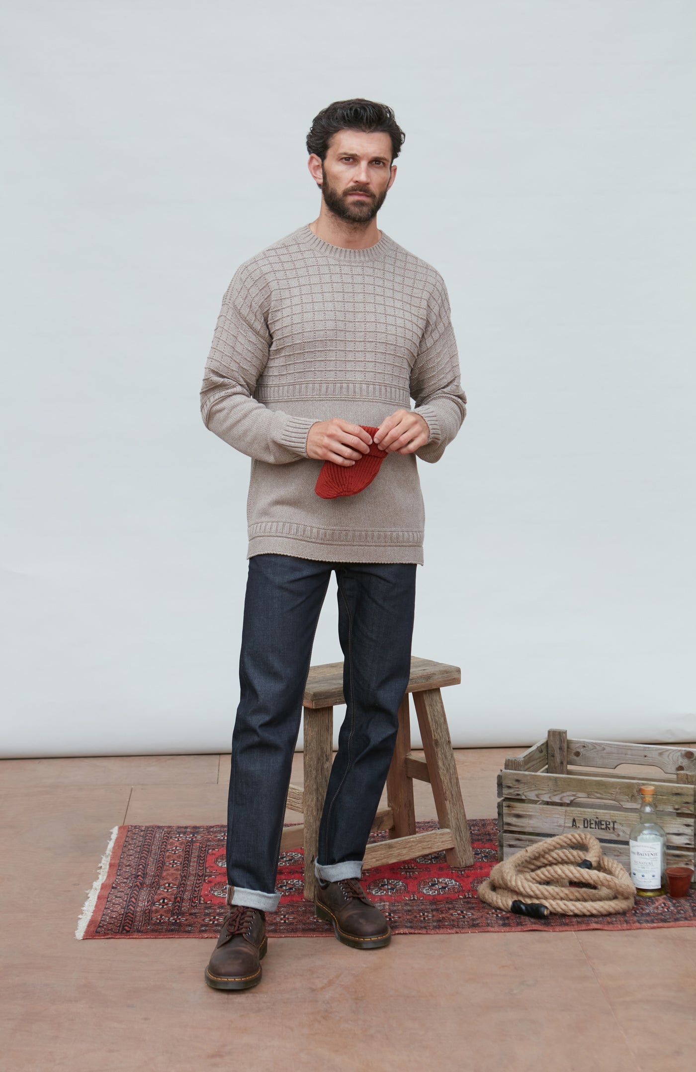 Man wearing a beige guernsey sweater and dark pants standing in a room with a wooden stool and rug.