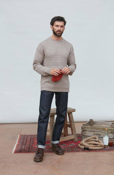 Man wearing a beige guernsey sweater and dark pants standing in a room with a wooden stool and rug.