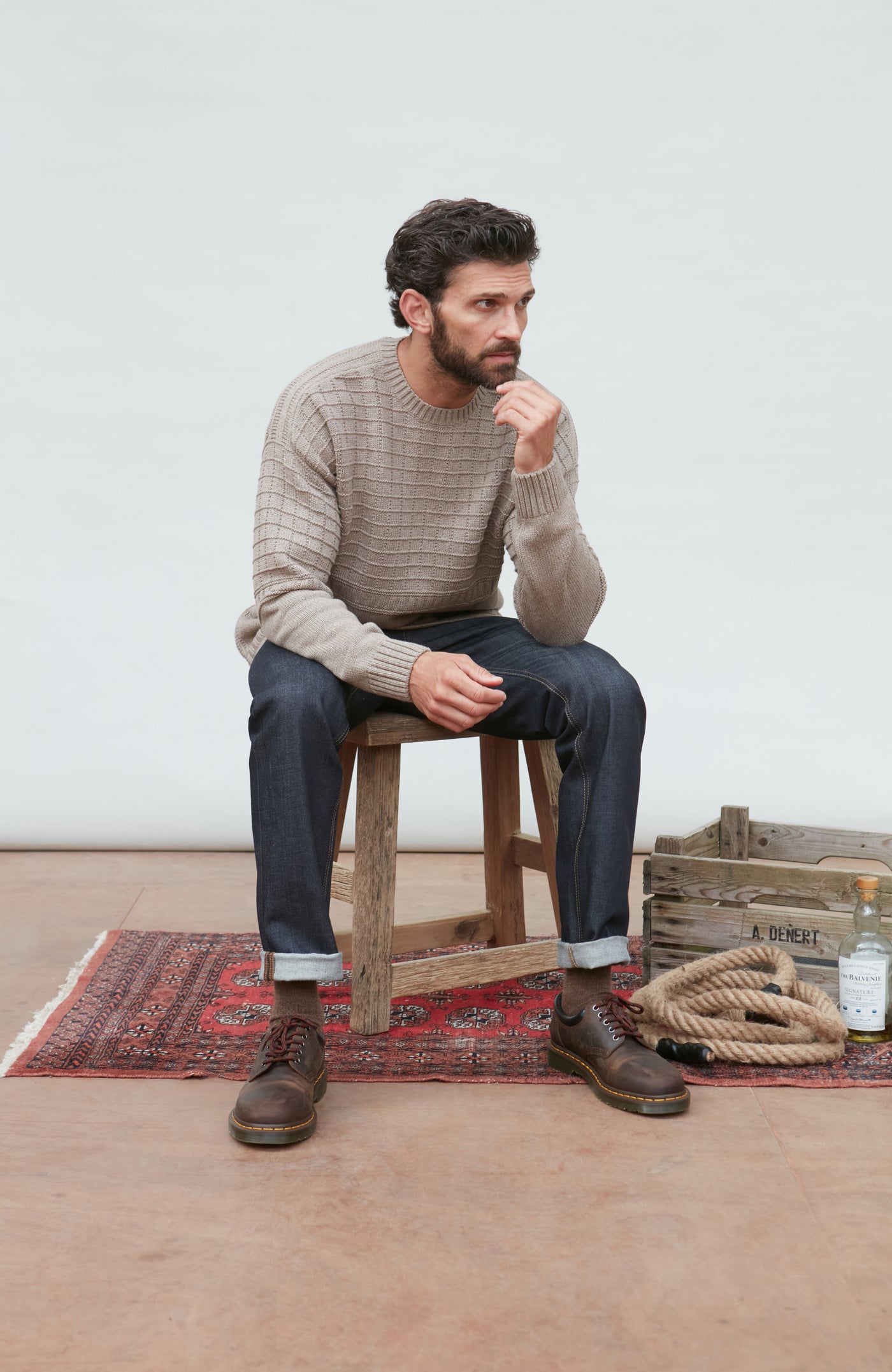 Man sitting on a wooden stool wearing a patterned sweater and jeans, with a white wall and wooden floor in the background.
