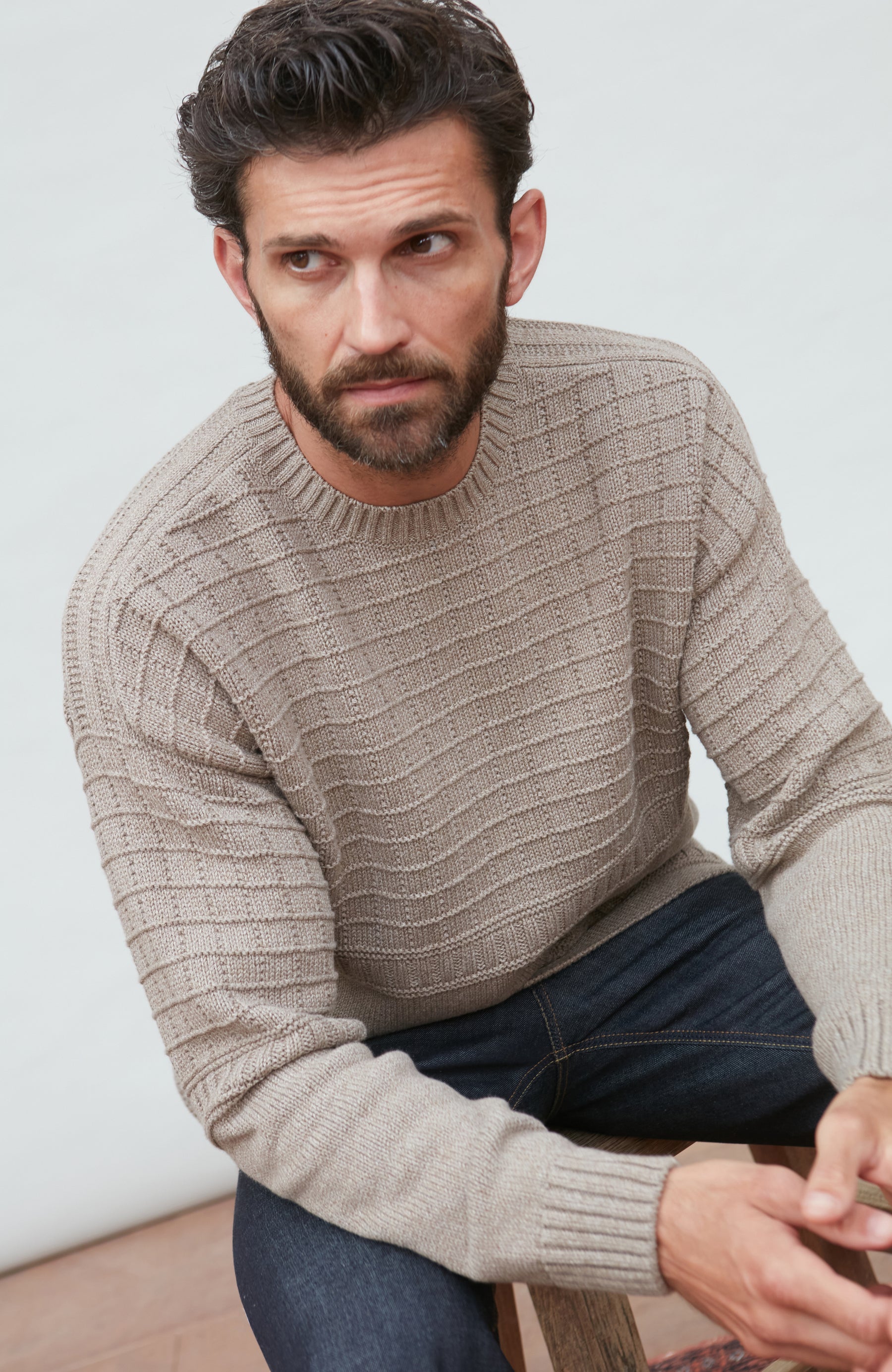 Man wearing a beige guernsey sweater sitting on a wooden stool against a light background