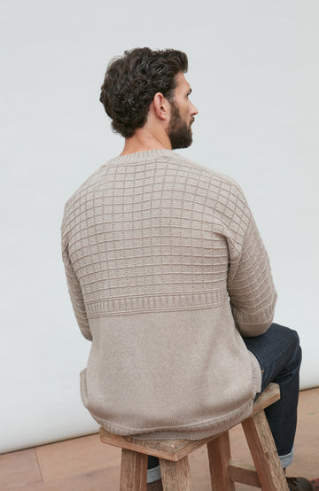 Man wearing a beige textured sweater sitting on a wooden stool against a white wall.