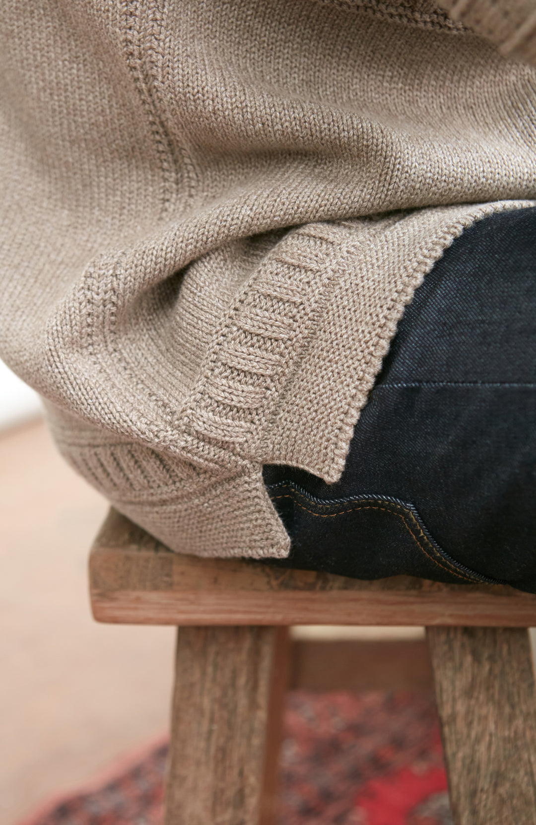 Person wearing a beige gansey sweater and dark jeans sitting on a wooden stool.