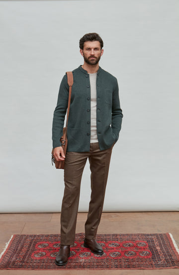 Man wearing a dark green mandarin collar jacket and brown pants standing on a red patterned rug with a white wall background.