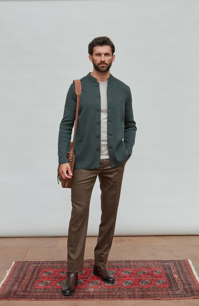 Man wearing a dark green mandarin collar jacket and brown pants standing on a red patterned rug with a white wall background.