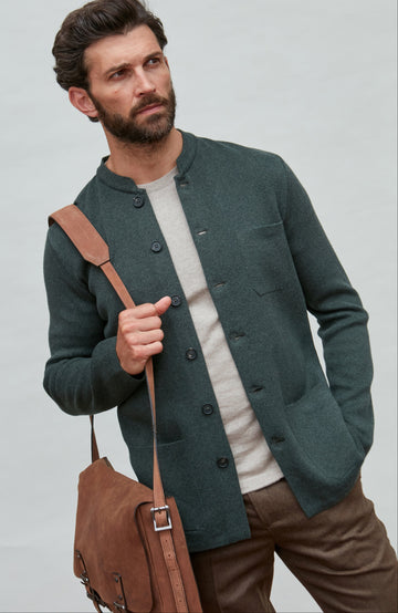 Man wearing a dark green mandarin collar jacket holding a brown leather bag against a light gray background