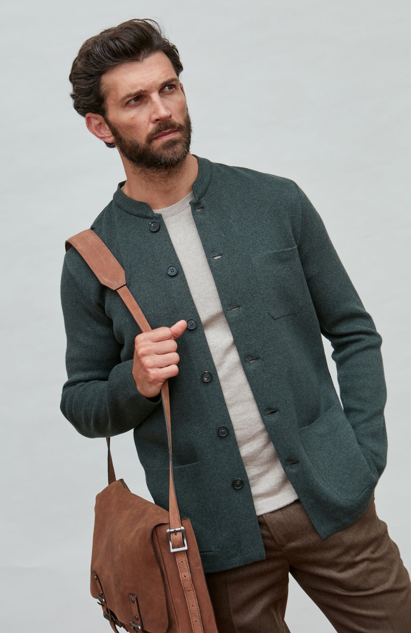 Man wearing a dark green mandarin collar jacket holding a brown leather bag against a light gray background