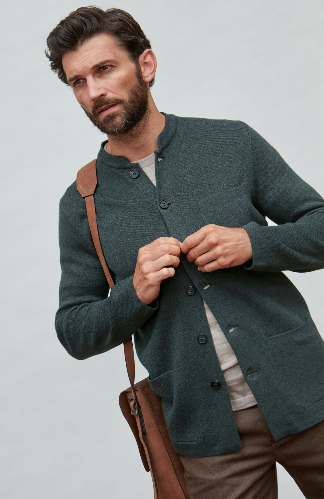 Man wearing a dark green mandarin collar jacket over a gray shirt, holding a brown leather bag against a light gray background.