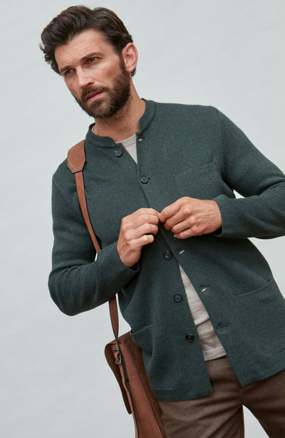 Man wearing a dark green mandarin collar jacket over a gray shirt, holding a brown leather bag against a light gray background.