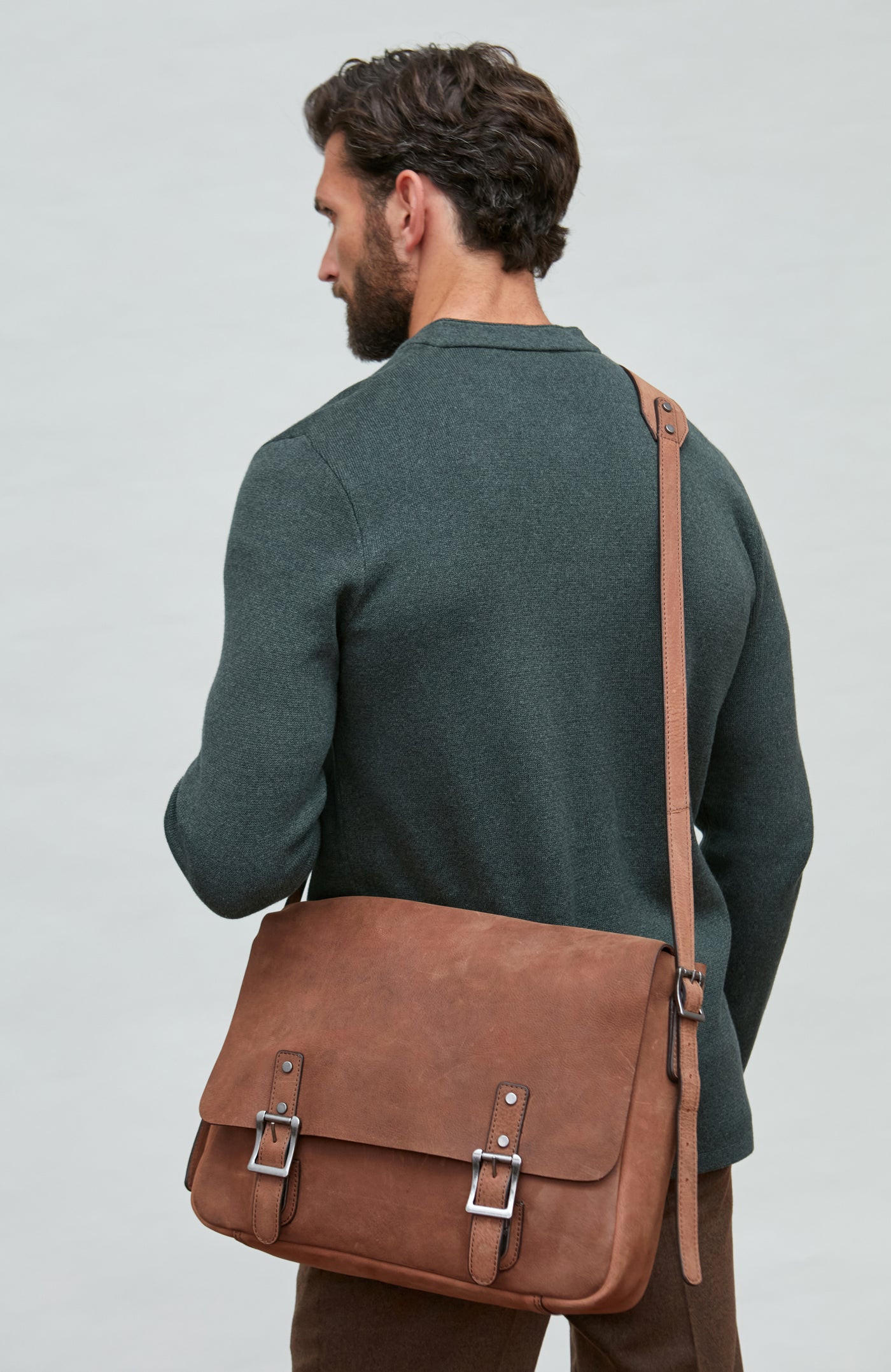 Man wearing a dark green mandarin collar jacket and brown leather satchel bag on a plain background