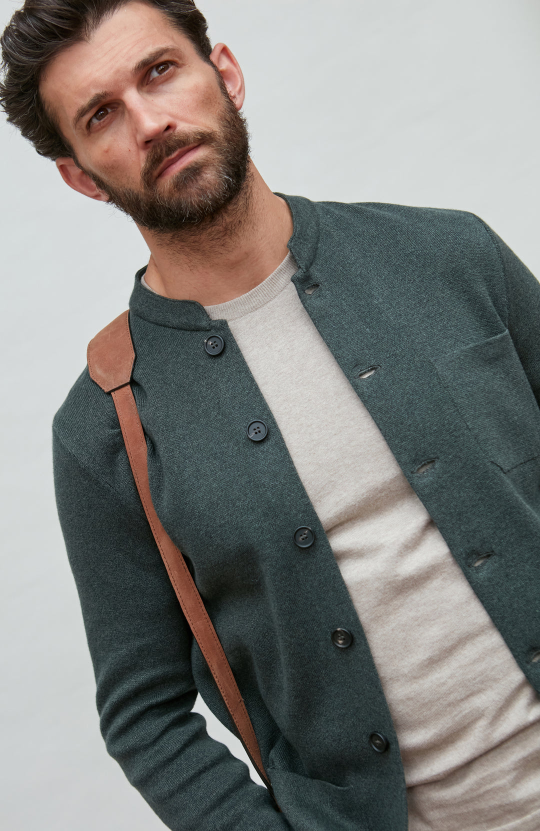 Man wearing a dark green mandarin collar  jacket over a white shirt with a brown strap over his shoulder on a light gray background