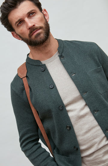 Man wearing a dark green mandarin collar  jacket over a white shirt with a brown strap over his shoulder on a light gray background