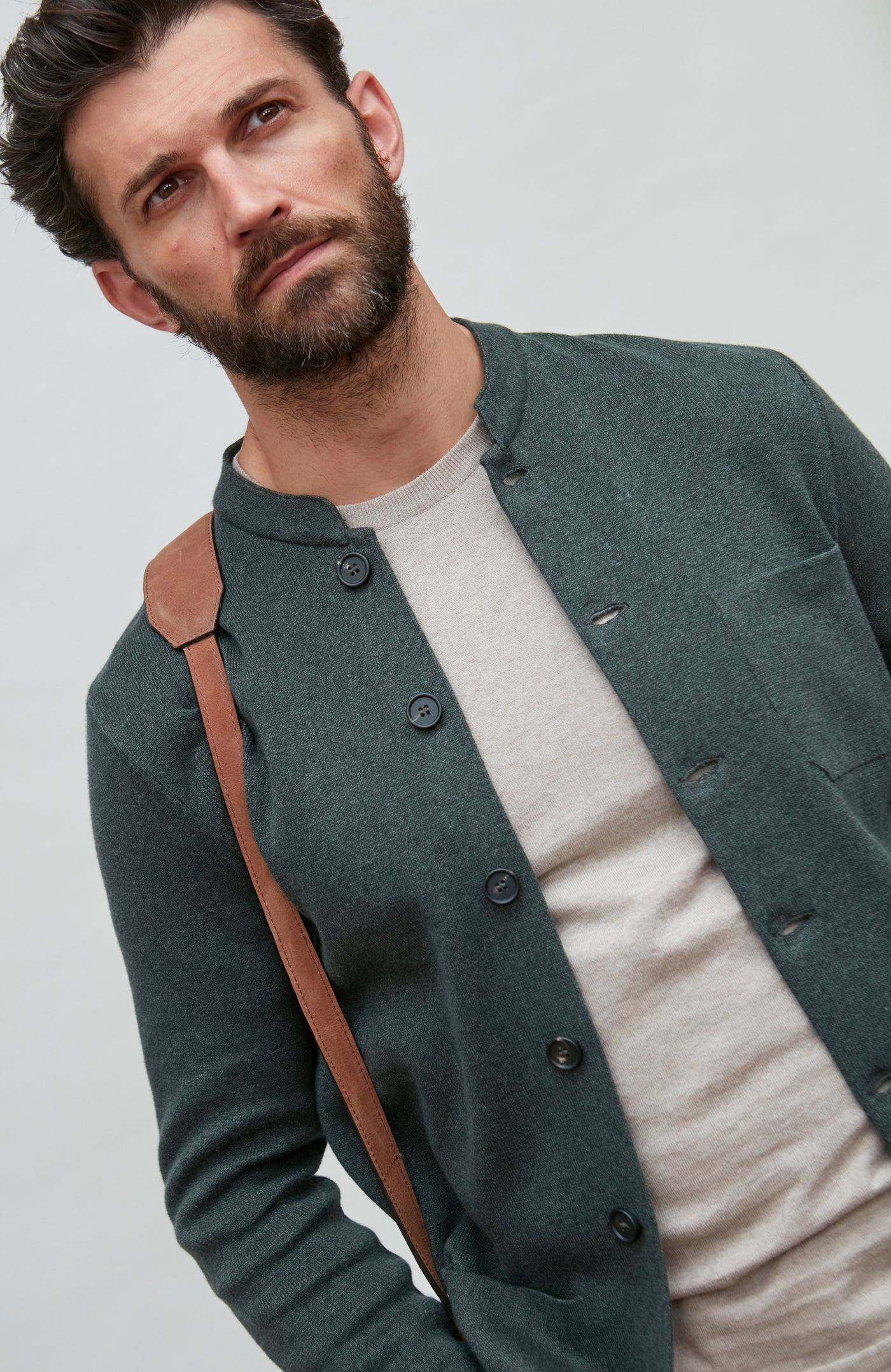 Man wearing a dark green mandarin collar  jacket over a white shirt with a brown strap over his shoulder on a light gray background