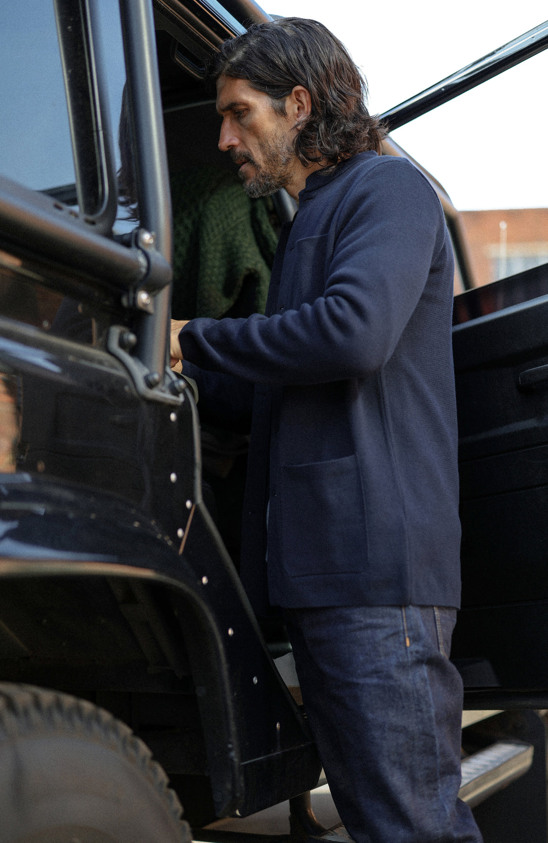 A man wearing a dark navy cotton nehru jacket with mandarin collar by Paul James Knitwear over a heathered blue-gray crewneck sweater, photographed in a real-world setting with a blurred background.