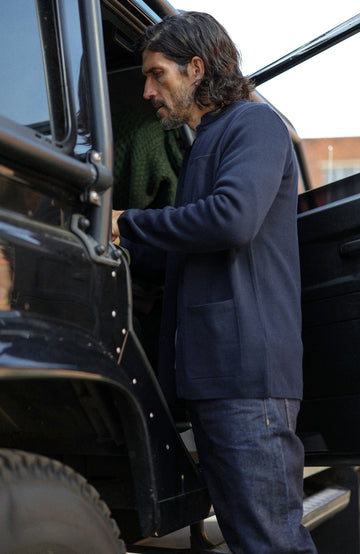 A man wearing a dark navy cotton nehru jacket with mandarin collar by Paul James Knitwear over a heathered blue-gray crewneck sweater, photographed in a real-world setting with a blurred background.