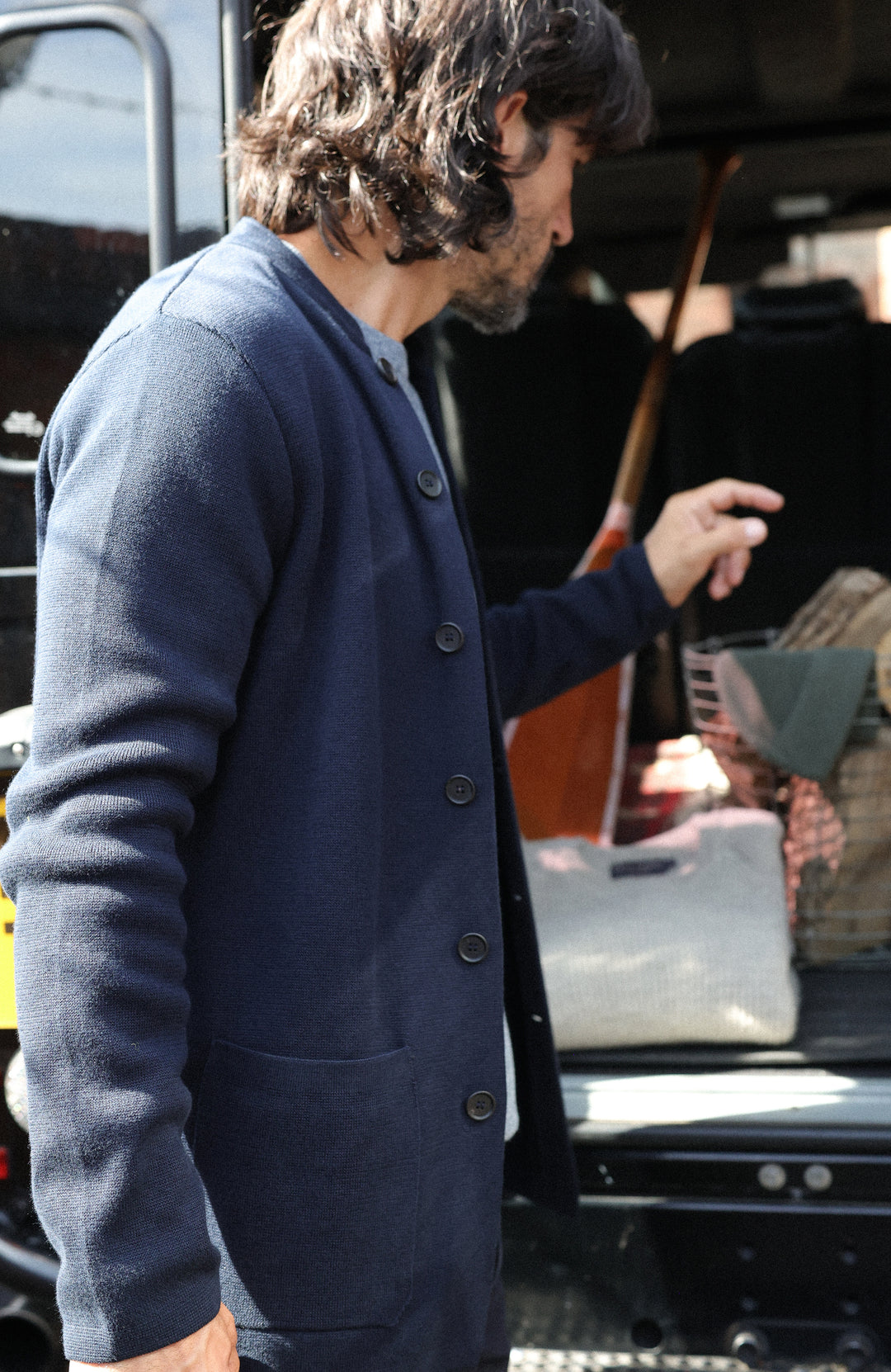 A man wearing a navy blue button-up nehru jacket stands next to an open vehicle trunk in daylight.