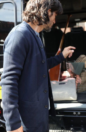 A man wearing a navy blue button-up nehru jacket stands next to an open vehicle trunk in daylight.