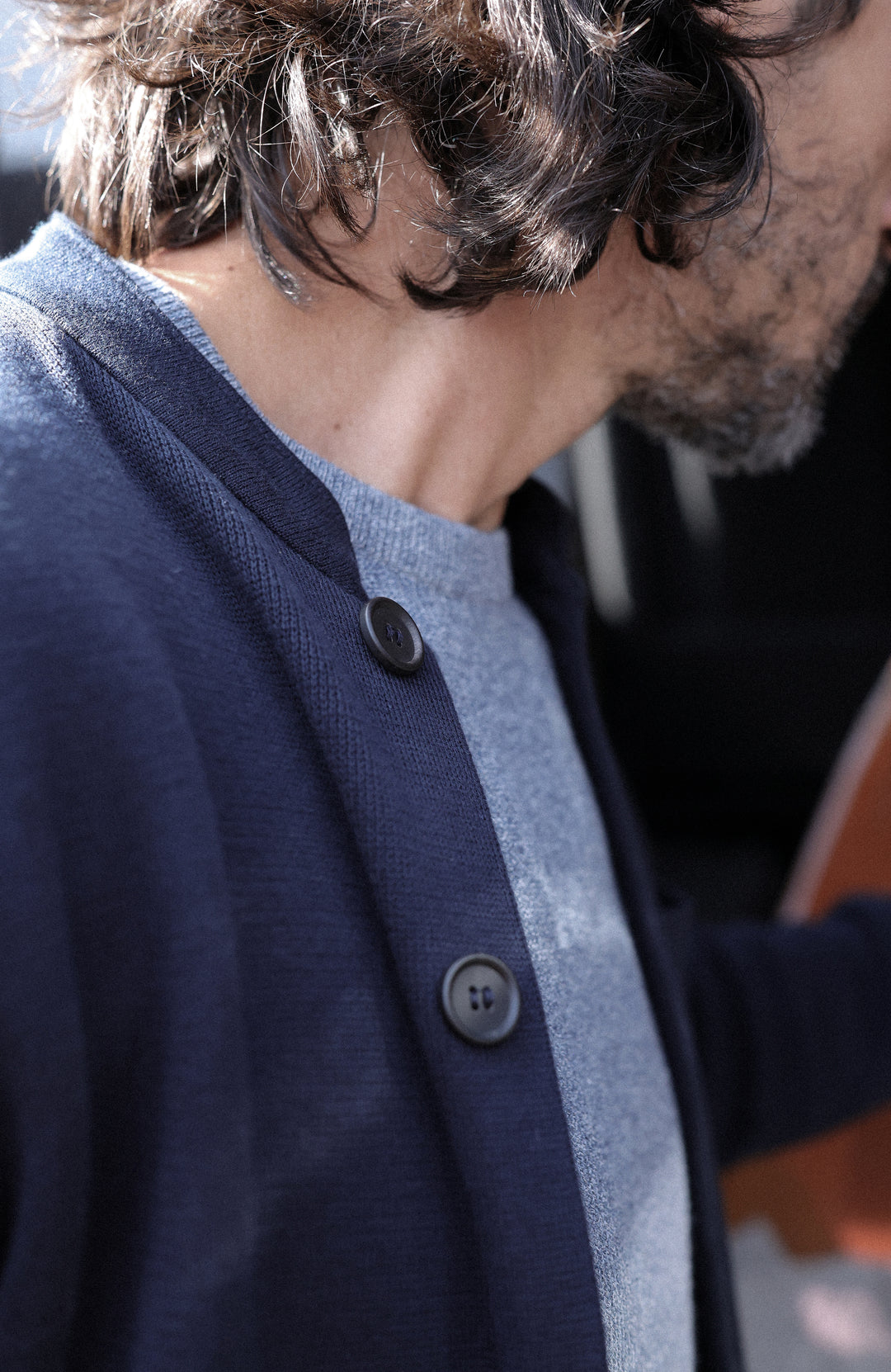 A man wearing a dark navy cotton nehru jacket with mandarin collar by Paul James Knitwear over a heathered blue-gray crewneck sweater, photographed in a real-world setting with a blurred background.