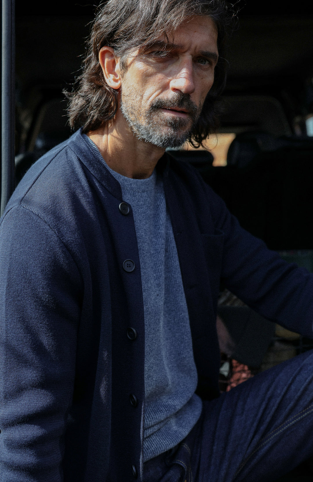 A man wearing a dark navy cotton nehru jacket with mandarin collar by Paul James Knitwear over a heathered blue-gray crewneck sweater, photographed in a real-world setting with a blurred background.
