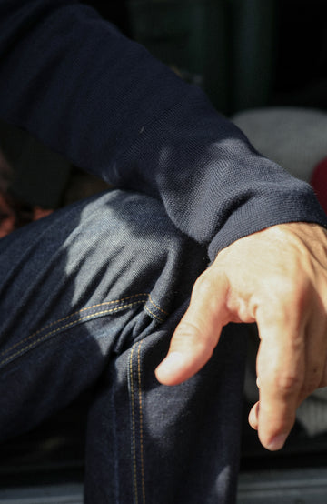 Close-up of a person wearing dark indigo denim jeans and a dark navy knit sweater, with sunlight casting shadows across the fabric and hand visible in the foreground.