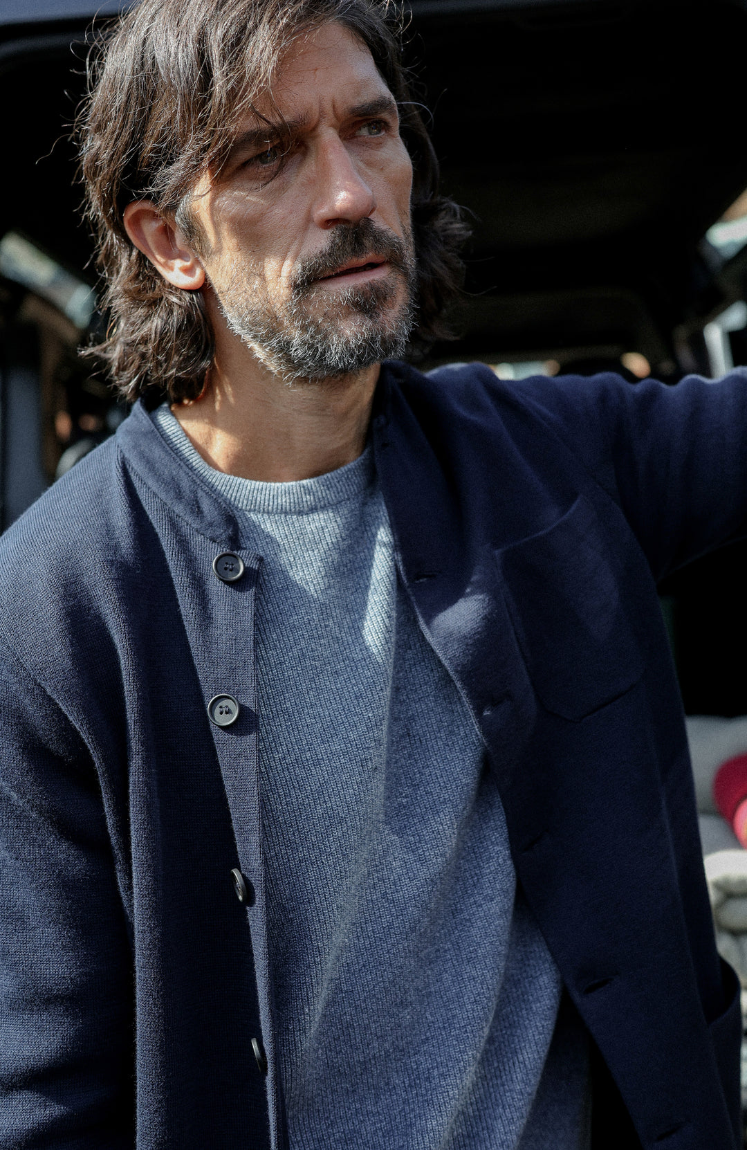 A man wearing a dark navy cotton nehru jacket with mandarin collar by Paul James Knitwear over a heathered blue-gray crewneck sweater, photographed in a real-world setting with a blurred background.