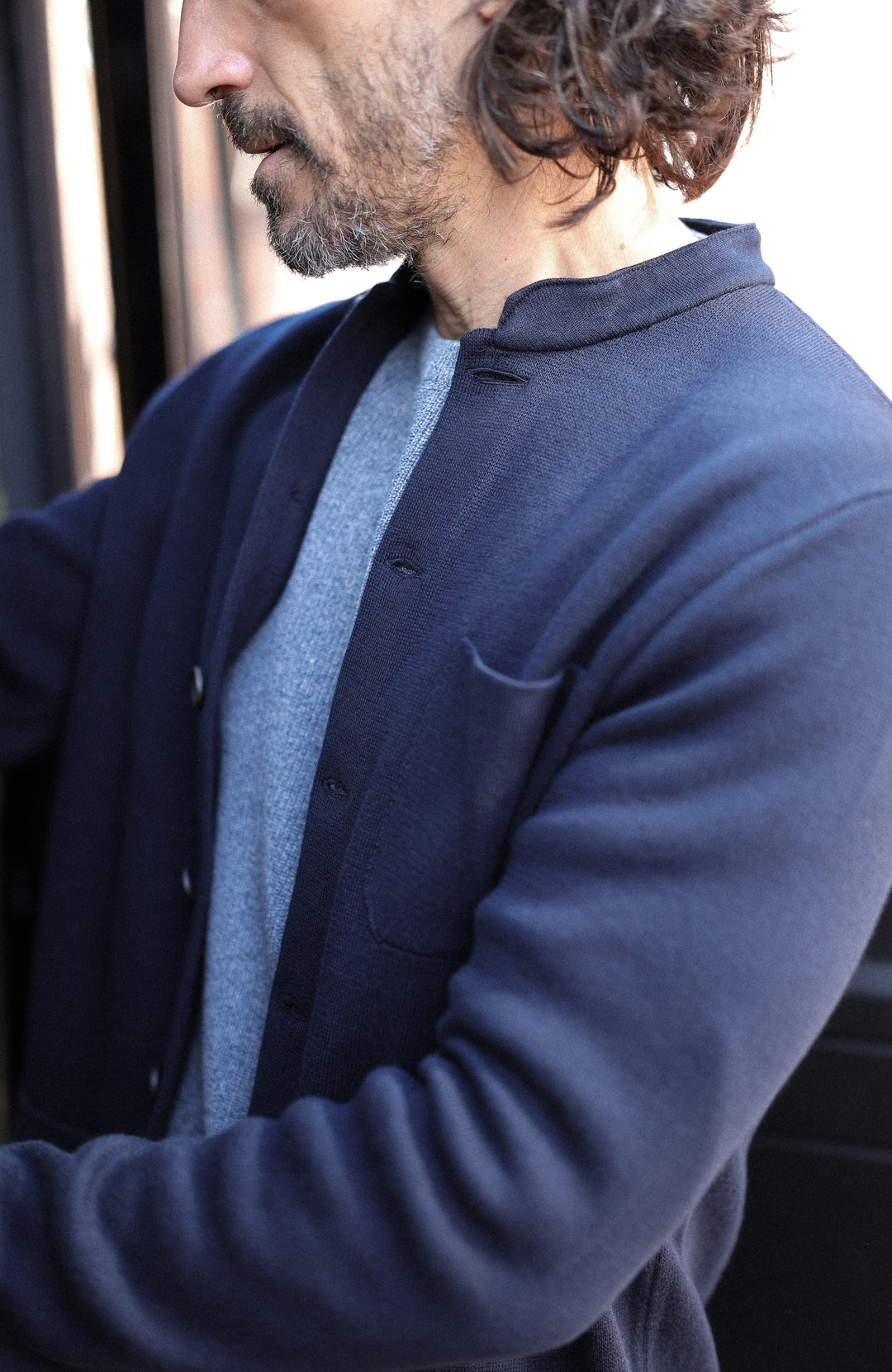 A close-up side profile of a man wearing a dark navy blue knit nehru jacket over a light gray sweater.