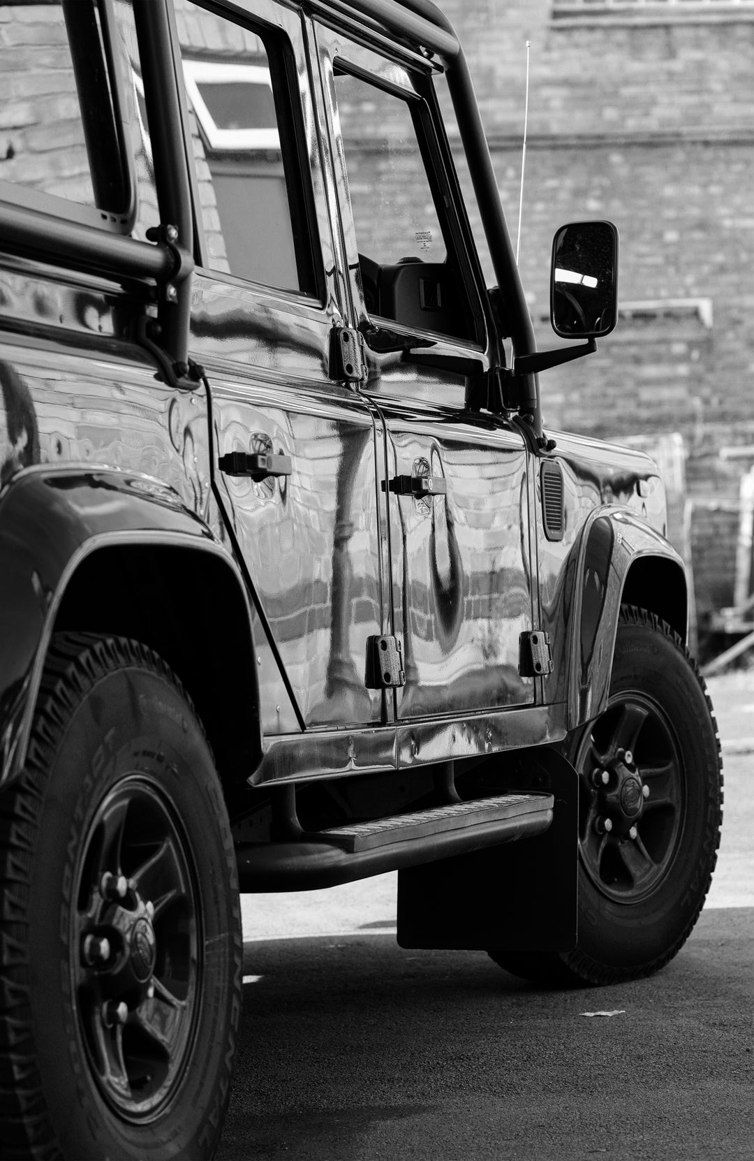 A black land rover defencer shown from a low side angle in a black-and-white photograph, with its front wheel, side door, mirror, and brick wall background visible.