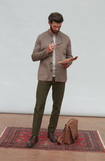 Man in a brown mandarin collar jacket  and green pants standing on a red patterned rug, holding glasses and a book.