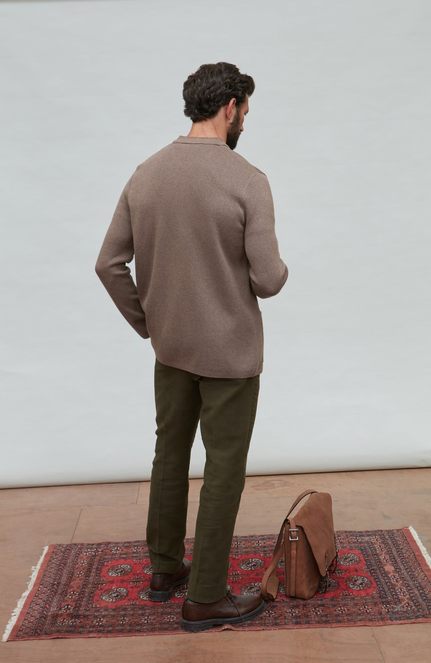 Man in a brown mandarin collar jacket  and green pants standing on a red patterned rug with a white wall background.