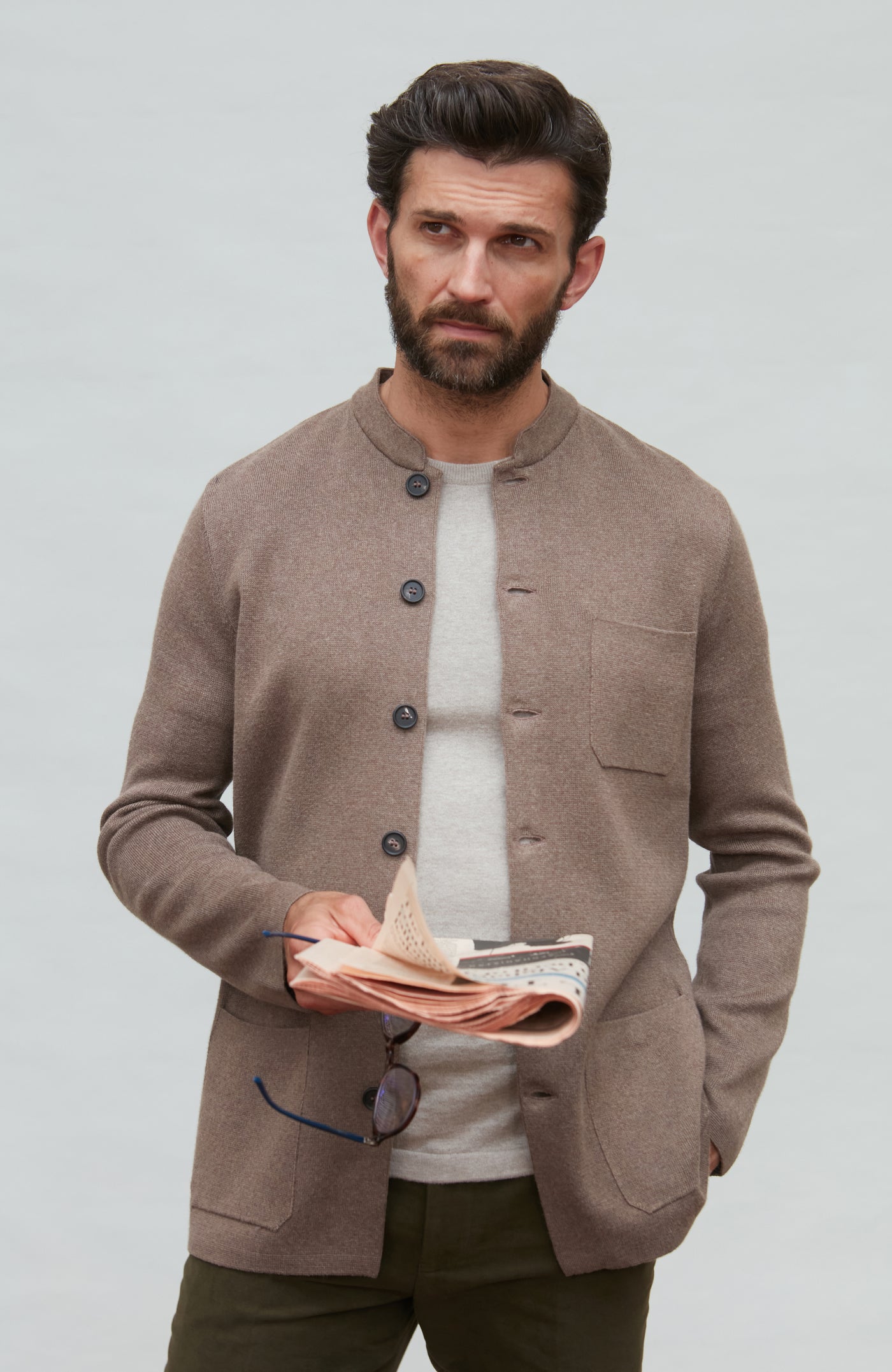 Man wearing a brown mandarin collar jacket over a white shirt, holding fabric against a plain background