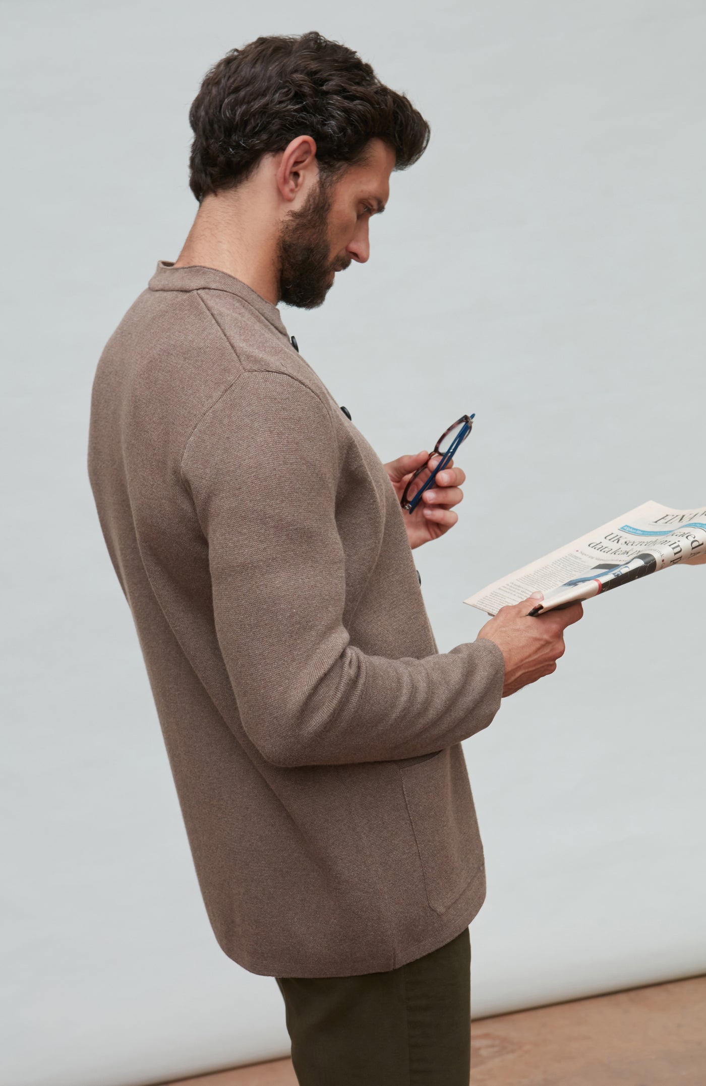 Man in a brown mandarin collar jacket  holding a phone and a book against a plain background